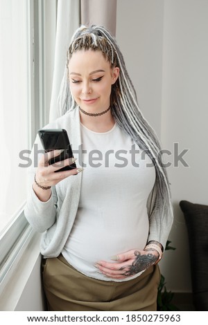Smiling attarctive young pregnant woman with dreadlocks standing at window, touching belly and reading news on social media