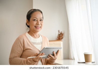 Smiling and attractive 60-year-old Asian woman in casual clothes sits in a minimal coffee shop with her digital tablet. - Powered by Shutterstock