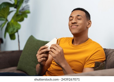 Smiling attractive African American man holding tasty sandwich, eating enjoying breakfast in morning while sitting on sofa at home. Young student having break, snack. Food concept - Powered by Shutterstock