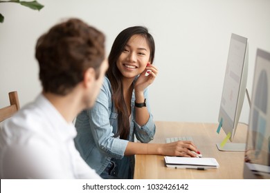 Smiling Attractive Asian Woman Talking To Male Colleague At Work Sharing Office Desk With Desktops, Friendly Multiracial Coworkers Interns Having Pleasant Fun Conversation At Workplace Together