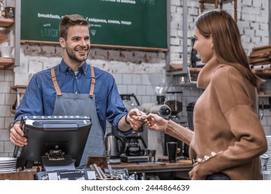 A smiling attractive barista waiter in apron stands behind a counter in a cafe, accepting credit card from a male customer to pay for her purchase order cup of coffee tea. The client making payment - Powered by Shutterstock