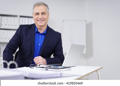 Smiling attractive businessman with a confident beaming smile sitting at his desk in the office working on paperwork