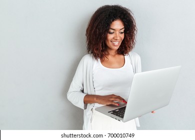 Smiling attractive casual african woman using laptop computer while standing isolated over white background