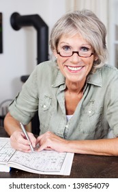 Smiling Attractive Elderly Woman Wearing Glasses Sitting At A Table Doing A Crossword Puzzle In A Book