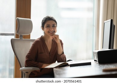 Smiling attractive Indian businesswoman sitting at desk in modern home office pose for camera feel confident, satisfied by business growth. Portrait of female freelancer, motivated student or employee