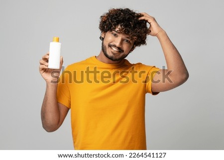 Smiling attractive Indian man holding a white bottle of moisturizing nourishing anti dandruff shampoo, looking at camera over isolated gray background. Hair care and medical treatment of hair loss
