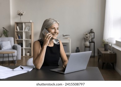 Smiling attractive mature woman sit at desk, work on laptop at home office, make mobile phone call, speak to client, giving consultation, lead formal pleasant conversation, remote job, communication - Powered by Shutterstock