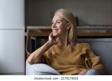 Smiling Attractive Middle-age Single Woman Daydreaming Staring Out Window Resting Alone Seated On Sofa In Studio Apartment. Homeowner Portrait, Rented Flat And Tenancy, Carefree Retired Life Concept