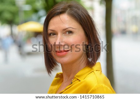 Smiling attractive middle-aged woman scrutinising the camera with a pensive expression in an urban square in a close up portrait