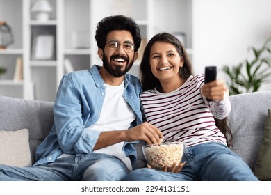 Smiling attractive millennial interracial spouses middle eastern man and insian woman in casual sitting on couch at home, hugging, watching tv, eating popcorn, enjoying favorite series or show