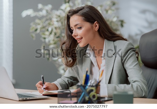 Smiling attractive pretty brunette woman in\
classic makeup wearing shirt jacket, sitting at desk in a company\
in front of a laptop, a secretary writes in a calendar\
appointments, important\
information
