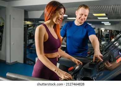 Smiling attractive red-haired girl in the gym with her trainer. Trainer adjust, press buttons at treadmill jogging track for pretty woman, gym customer, sportive lifestyle - Powered by Shutterstock