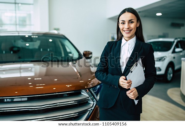 Smiling attractive\
saleswoman holding documents while looking at camera at new car\
showroom or dealership