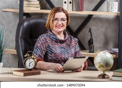 Smiling attractive senior businesswoman wearing glasses, working on tablet in modern office. Happy aged teacher and successful woman company boss.