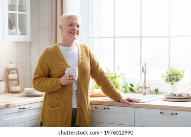 Smiling attractive senior lady with mug, drinking fresh coffee or tea at kitchen alone. Happy grandmother in casual outfit holding cup, standing in the cozy kitchen, looking at copy space - Powered by Shutterstock