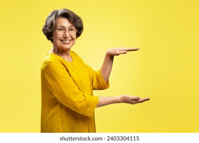 Smiling attractive senior woman holding hands with empty space looking at camera isolated on yellow background. Shopping, store, advertisement concept 