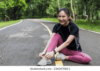 Smiling attractive sportswoman sitting on road and tiring shoeless in green park - Shutterstock ID 2360470811