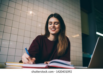 Smiling attractive student programmer doing homework checking information using modern computer, skilled happy female  writing ideas to notebook sitting in college campus and enjoying learning time