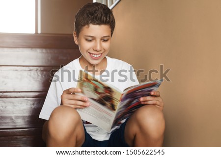 Smiling attractive teen boy sitting at home on steps and reading magazine. Cheerful guy in white t-shirt is sitting at home, reading comic book. Schoolboy is reading paper book while resting home.
