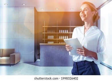 Smiling Attractive Woman Holding Tablet Device And Waiting For Clients In Reception Area Of Modern Luxury Spa Wellness Center To Sign In Membership Contract. Towels, Soaps And Perfumes Are On Shelves