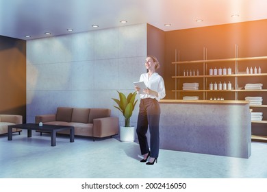 Smiling Attractive Woman Holding Tablet Device And Waiting For Clients In Reception Area Of Modern Luxury Spa Wellness Center To Sign In Membership Contract. Towels, Soaps And Perfumes On Shelves