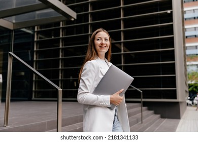 Smiling Attractive Woman With Long Straight Hair Wearing White Jacket Walking In Business District On Background Go Modern Building With Laptop.