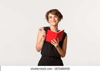 Smiling attractive woman taking notes in planner, prepare working schedule in notebook, standing over white background