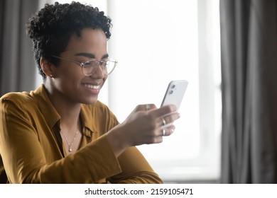 Smiling attractive young African woman looks at cellphone screen seated at table indoor. Read sms, scroll news feed spend free time on internet, use mobile application, modern tech usage, fun concept - Shutterstock ID 2159105471