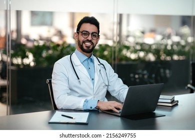 Smiling attractive young arab man doctor with beard in glasses and white coat, at workplace work with laptop in office interior. Consultation and diagnostics remotely, modern gadget and new normal