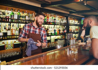 Smiling Attractive Young Barman Wiping Glasses And Talking To African American Young Man In Bar