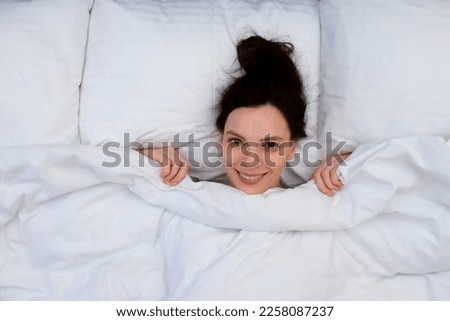 Smiling attractive young brunette woman is hiding under the blanket laying in bed in the morning on white linen. The concept of a lazy morning and unwillingness to get up. Hiding from reality.