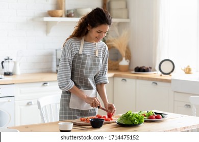 Smiling attractive young curly woman wearing apron, chopping fresh colorful vegetables for salad on wooden board. Happy housewife preparing healthy food for vegan dinner alone at modern kitchen. - Powered by Shutterstock