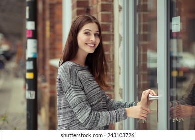 Smiling Attractive Young Female Student Entering A Commercial Building Looking At The Camera As She Pushes Open The Glass Door