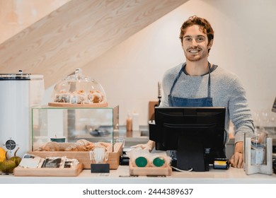 Smiling attractive young waiter standing at cafe counter in small cafeteria. Male cafeteria staff vendor seller selling food and drinks to customers. Customer service business concept - Powered by Shutterstock