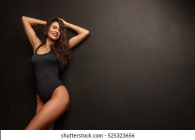 Smiling attractive young woman with sexy shapely forms, loose wavy hair, red lipstick and slim body is posing in the black underwear, studio photoshoot, dark background