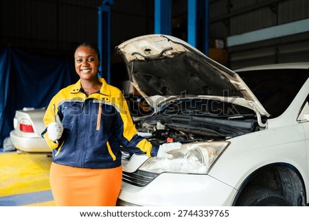 Smiling auto mechanic in a workshop stands beside a white car with the hood open, wearing gloves and a work jacket, ready for vehicle maintenance