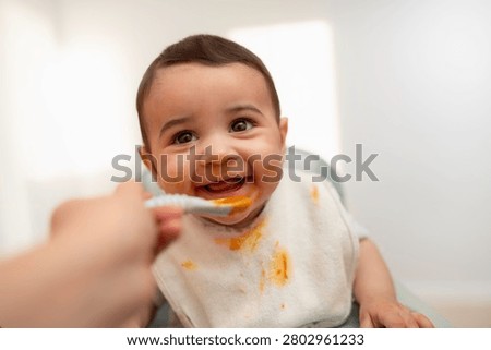 Smiling baby enjoying first solid foods while being fed with a spoon. Cute infant wearing a bib during mealtime at home, illustrating baby-led weaning, complementary feeding, and healthy child development.