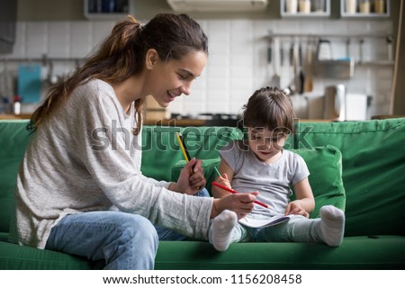 Smiling baby sitter and preschool kid girl drawing with colored pencils sitting on sofa together, single mother and child daughter playing having fun, creative family activities at home concept