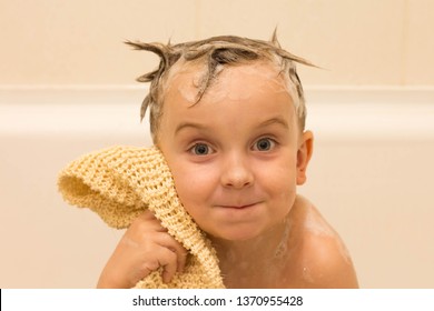 Smiling Baby Is Sitting In Bathtub With Shampoo Foam On Hair And Washcloth In Hand, White Background, Isolated