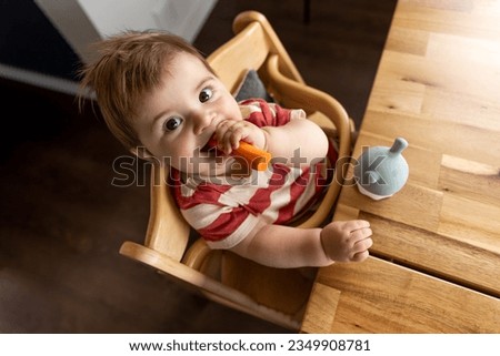 Smiling baby sitting in a baby chair and eating vegetables by own hand