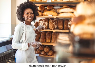 Smiling baker woman standing with fresh bread at bakery. Happy african woman standing in her bake shop and looking at camera. Satisfied baker with breads in background. Smiling woman at bakery shop - Shutterstock ID 1804460734