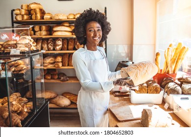 Smiling baker woman standing with fresh bread at bakery. Happy african woman standing in her bake shop and looking at camera. Satisfied baker with breads in background. Beautiful  woman at bakery shop - Powered by Shutterstock