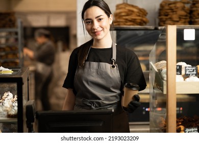 Smiling Baker Woman Standing With Fresh Bread And Pastry At Bakery. Young Woman In Her Bake Shop And Looking At Camera. Baker With Breads In Background. Girl Owner Bakery Shop
