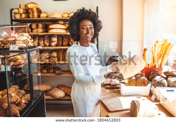 Smiling baker woman standing with fresh bread at\
bakery. Happy african woman standing in her bake shop and looking\
at camera. Satisfied baker with breads in background. Beautiful \
woman at bakery shop