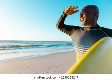 Smiling bald african american senior man shielding eyes and carrying surfboard at beach against sky. Copy space, water sports, recreation, retirement, unaltered, vacation, enjoyment, nature, sunset. - Powered by Shutterstock