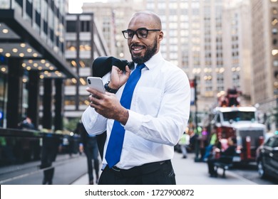 Smiling bald bearded African American executive in white shirt with blue tie and eyeglasses holding jacket on shoulder and texting message on downtown street 