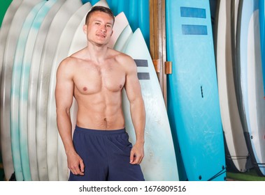 Smiling bare-chested guy surfer posing with surfboard on shoulders