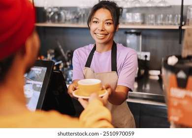 Smiling barista handing a cup of coffee to a customer in a cafe. The barista is wearing a purple shirt and a beige apron - Powered by Shutterstock