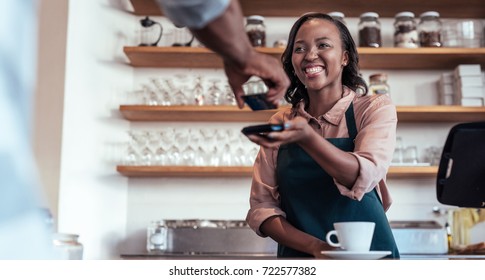 Smiling barista using nfs technology to help a customer pay for a purchase with their bank card in a cafe 