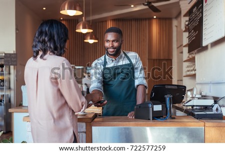 Smiling barista using nfs technology to help a customer pay for a purchase with their smartphone in a cafe 